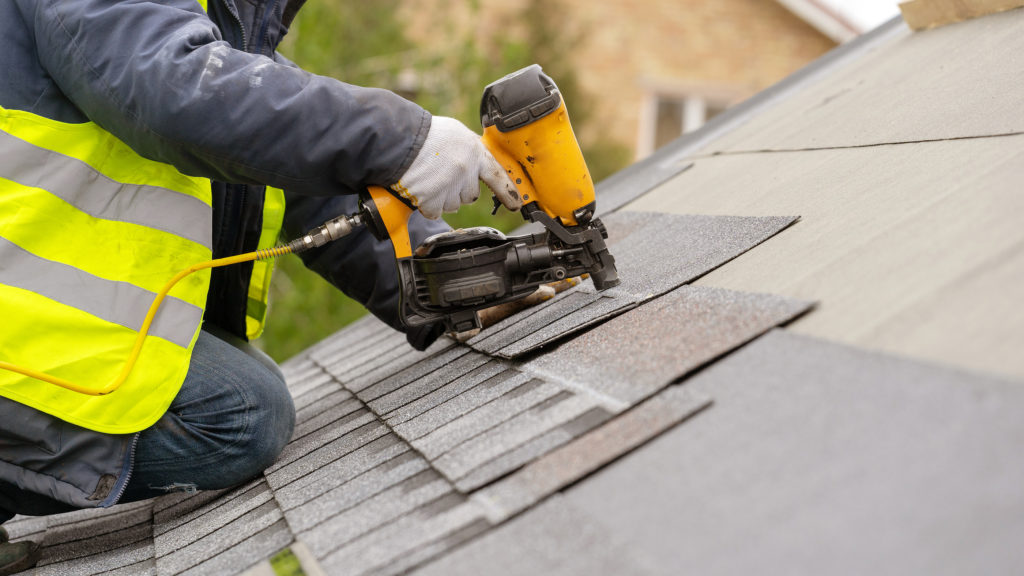 A roofer installing asphalt shingles with a nail gun.